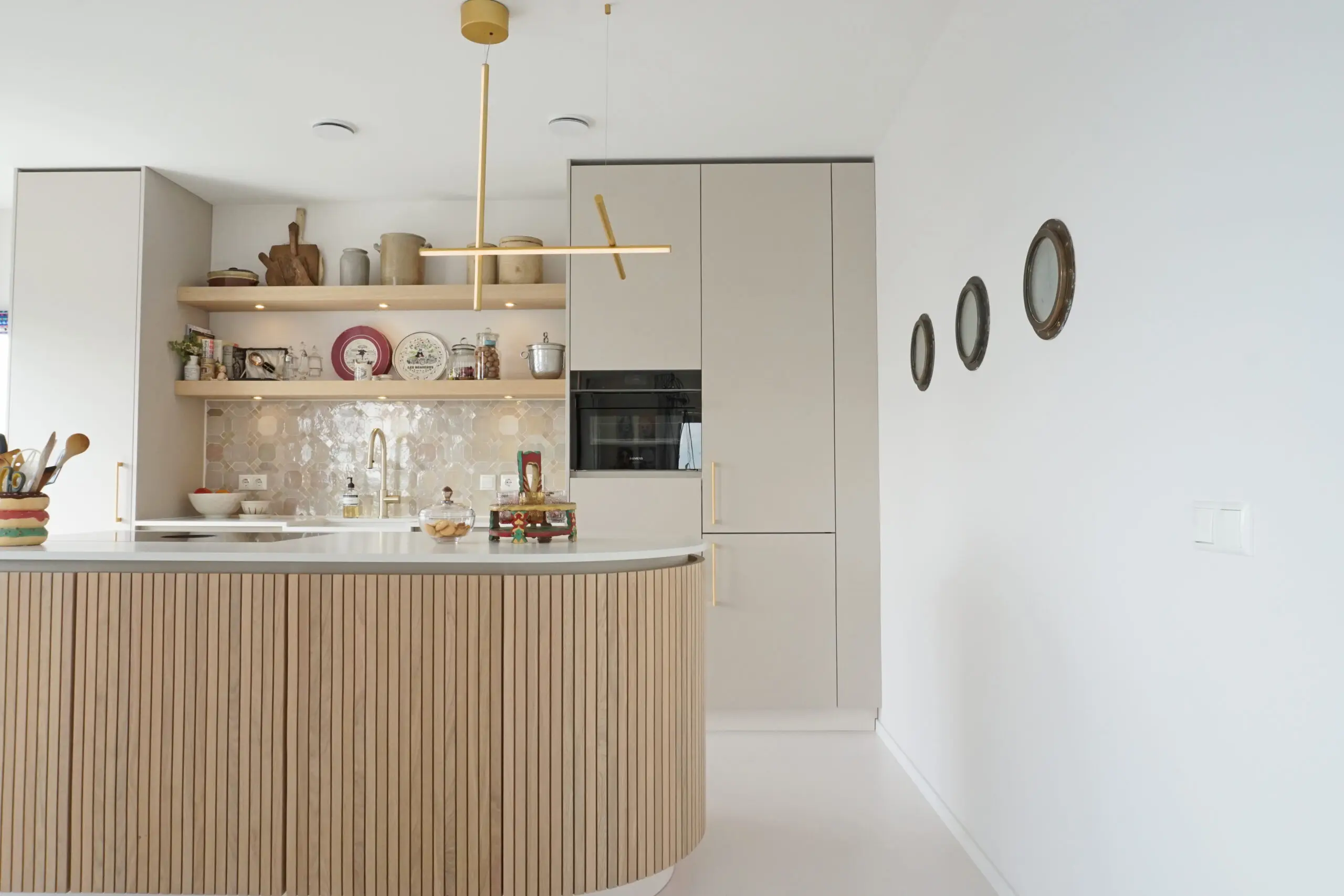 Warme keuken met gebogen houten kookeiland, greige kasten, open houten planken en een messing hanglamp. Interieurproject Appartement Maashaven Rotterdam