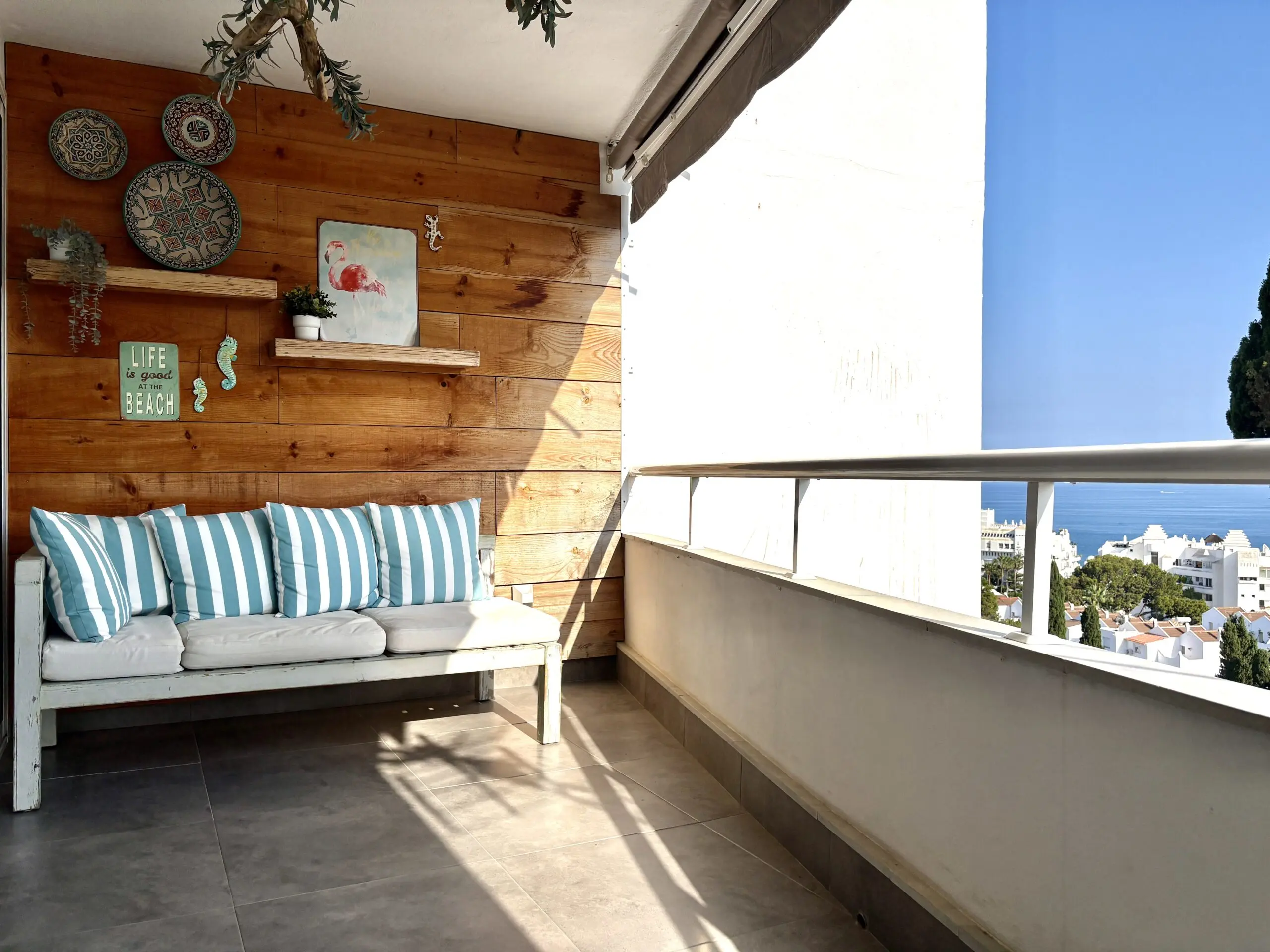"Zonnig balkon met grijze tegelvloer en een houten wandbekleding. Tegen de houten wand een wit geschilderde houten bank met grijs-wit gestreepte kussens. Op een houten wandplank staan een wit plantenpotje en een flamingo-bordje, omgeven door decoratieve Marokkaanse borden, zeepaardje-ornamenten en een tekstbord met 'Life is good at the Beach'. Rechts een uitgerold zonnescherm en een wit hekwerk met uitzicht over een mediterraan stedelijk landschap met cipressen en de blauwe zee in de verte onder een heldere blauwe hemel." Interieurproject Málaga